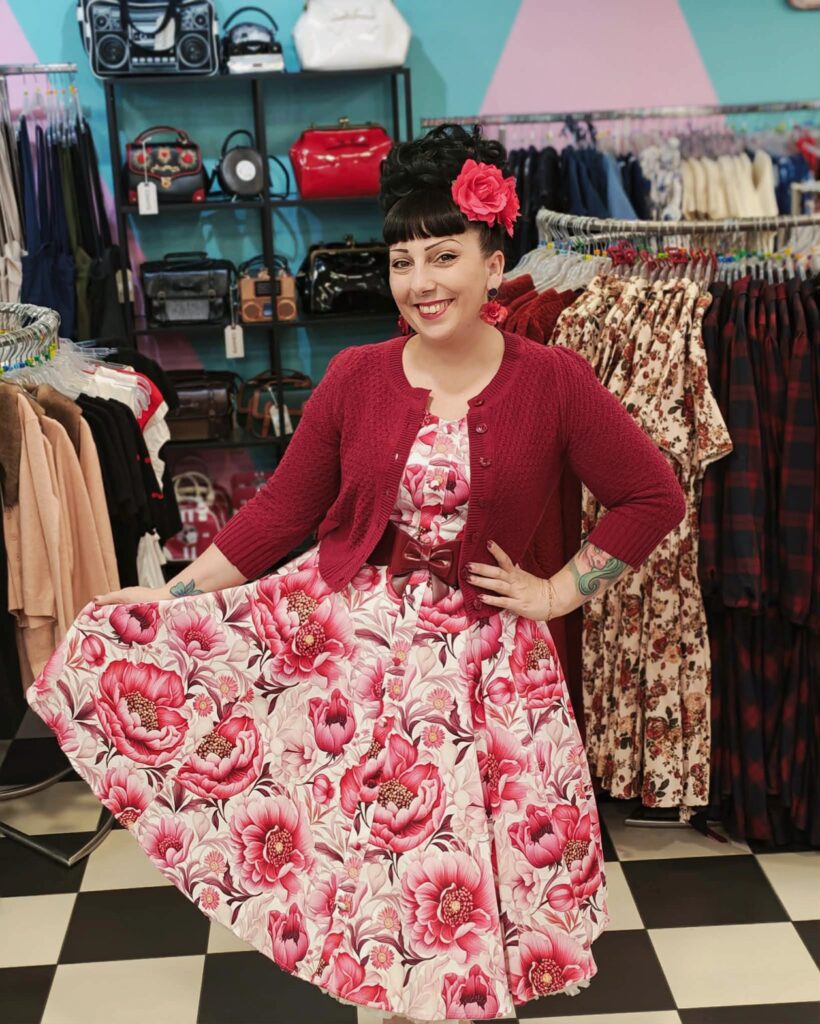 Femme portant une robe pin-up à fleurs rose fuchsia et bordeaux sur fond blanc avec un cardigan en crochet écru - Sweet Candy Shop